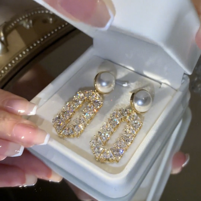 Gold and pearl earrings in a jewelry box held by a hand.