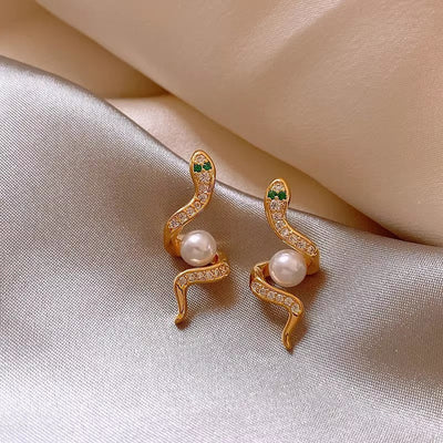 Gold snake-shaped earrings with pearl centers on a soft fabric background