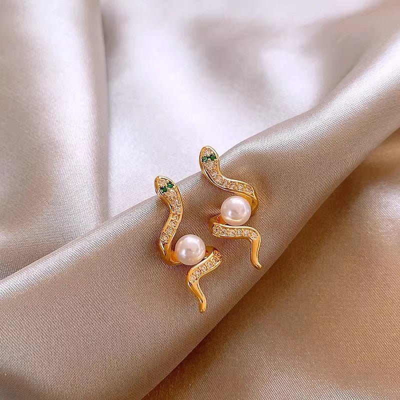 Gold snake-shaped earrings with pearl accents on a beige fabric background