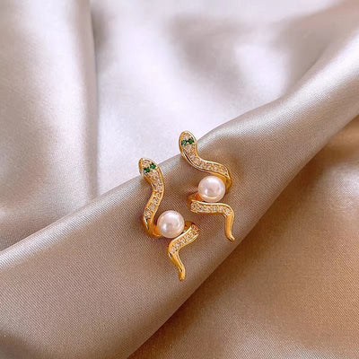 Gold snake-shaped earrings with pearl accents on a beige fabric background