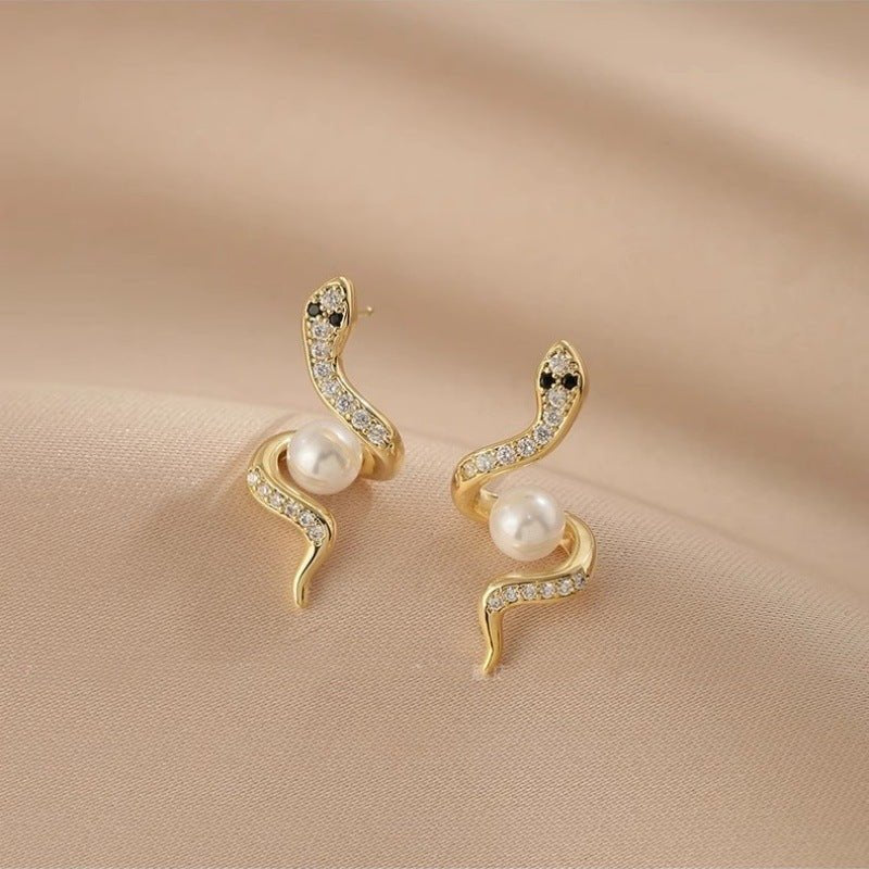 Gold snake-shaped earrings with pearls on a beige fabric background